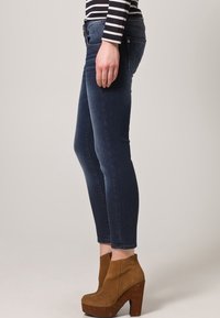 Osoba v tmavě modrých slim-fit džínech, pruhované košili s dlouhým rukávem a hnědých semišových vysokých botách na kotníky, stojící bokem.