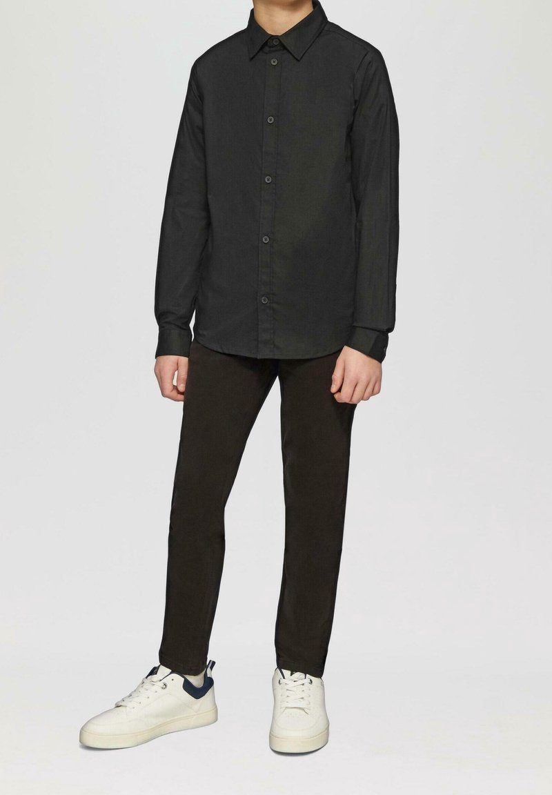 Personne portant une chemise noire à manches longues boutonnée, un pantalon noir et des baskets blanches avec des accents sombres au talon, debout sur un fond uni.