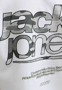 Κοντινό πλάνο του κειμένου «Jack Jones» με σχέδιο παραλλαγής σε μαύρο και γκρι πάνω σε λευκό ύφασμα, με μικρότερα εκτυπωμένα στοιχεία μάρκας και πνευματικών δικαιωμάτων από κάτω.