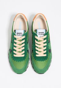 Zielone trampki wykonane z mieszaniny materiałów, z falistymi zielonymi akcentami, czarnymi detalami oraz beżowymi sznurowadłami. Pomarańczowa pętla na pięcie i widoczna logo na wkładce.