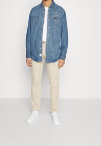 Persona che indossa una camicia di jeans blu con bottoni sopra una t-shirt bianca, pantaloni cargo beige e scarpe da ginnastica bianche, in piedi contro uno sfondo semplice.