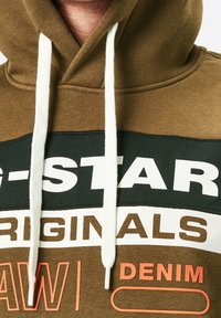 Sudadera marrón con cordones blancos, que presenta patrones de texto en negrita en blanco y negro, además de acentos naranjas sobre un fondo texturizado.