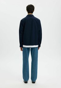 Chemise bleu marine à coupe décontractée, avec des détails à boutons et des manches longues, portée sur une chemise blanche ; associée à un pantalon en denim bleu à jambes larges.