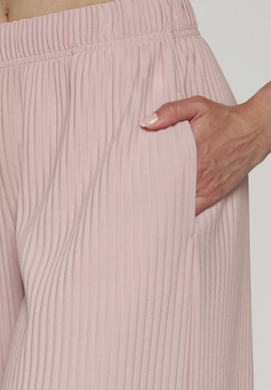 Hand with light pink polished nails resting inside the pocket of pale pink vertically pleated elastic waistband pants.