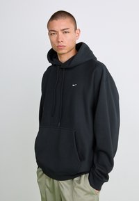 Ung mand med buzz cut iført sort Nike hoodie og lys beige bukser, stående med hænderne i lommerne mod en ensfarvet grå baggrund.