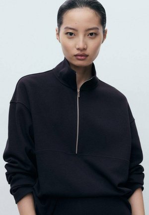 Ragazza giovane con i capelli slicked-back che indossa una felpa nera con zip e maniche arrotolate, sfondo neutro.