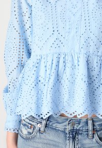 Blusa azzurro chiaro con ricamo ad occhio, orlo scallop e maniche lunghe con dettagli in pizzo, abbinata a jeans blu scoloriti.