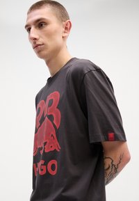Dark gray cotton T-shirt featuring a large red graphic design of a bull and text. Short sleeves, relaxed fit, and branded tag on sleeve.