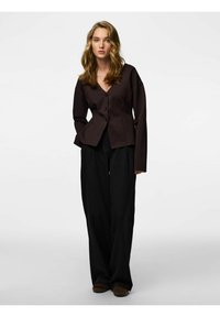 Hnědá bunda na knoflíky s výstřihem do V, kombinovaná s černými kalhotami s bílými proužky a širokými nohavicemi. Model stojí vzpřímeně s rukama v kapse.