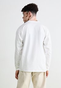T-shirt blanc en coton à manches longues avec un col rond et une coupe décontractée, vu de dos. Porté avec un pantalon beige clair.