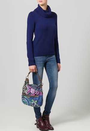Mujer con suéter de cuello alto azul, vaqueros azules y botines burdeos sosteniendo un bolso de mano multicolor con motivos de mariposas.