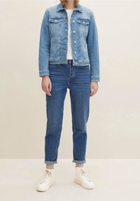 Persona vistiendo una chaqueta de mezclilla azul claro, camiseta blanca, jeans oscuros arremangados, calcetines beige y zapatillas blancas de pie contra un fondo liso.