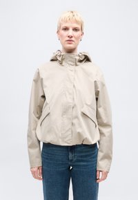 Άτομο με κοντά ξανθά μαλλιά που φοράει μια μπεζ ζακέτα με κουκούλα και μπλε τζιν, στέκεται μπροστά σε ένα απλό λευκό φόντο.