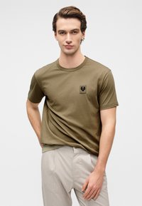 Πουκαμίσα από βαμβάκι σε ελιά πράσινο χρώμα με στρογγυλή λαιμόκοψη και κοντά μανίκια, με μια μικρή μαύρη ετικέτα λογότυπου στο αριστερό στήθος.