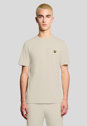 Jeune homme aux cheveux courts blonds portant un t-shirt beige et un pantalon assorti, avec un petit logo jaune et noir sur la poitrine.
