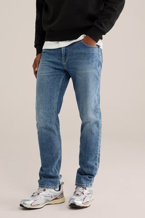 Person trägt blaue Jeans und silberne Sneaker mit roten Details, steht mit einer Hand in der Tasche vor einem schlichten Hintergrund.