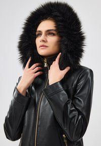 Giubbotto in pelle nera con cappuccio in pelliccia sintetica. Presenta una cerniera dorata e dettagli discreti delle tasche. Texture liscia e design su misura.