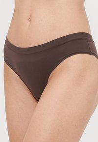 Smeđi bikini donji dio napravljen od glatkog materijala, s niskim strukom, čistim rubovima i minimalističkim stilom.