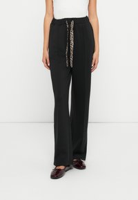 Zwarte, wijde broek met een koordsluiting in de taille, voorzien van een luipaardprint detail. De stof lijkt zacht en soepel. Platte, glanzende bordeauxrode schoenen.