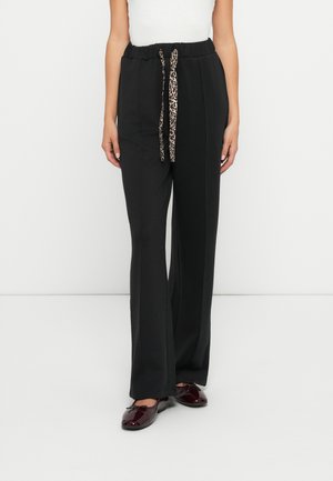 Pantalons noirs à jambe large avec une taille à cordon et un détail en imprimé léopard. Le tissu semble doux et lisse. Chaussures bordeaux plates et brillantes.