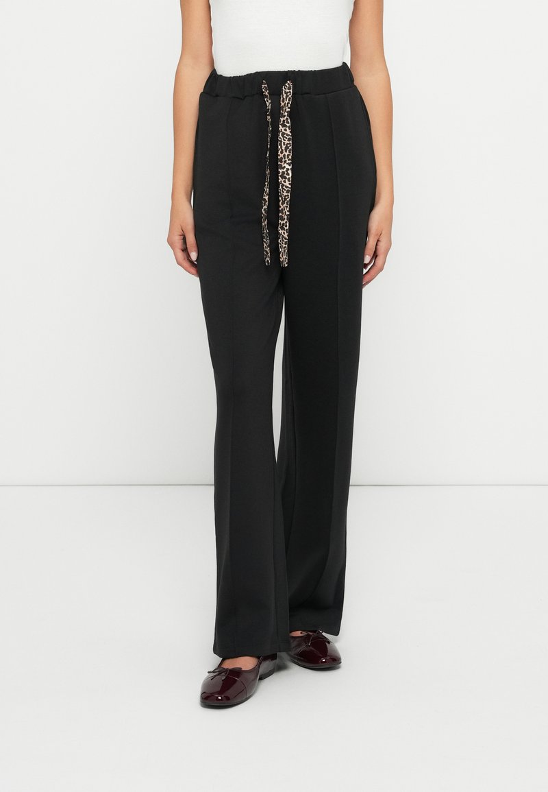 Zwarte, wijde broek met een koordsluiting in de taille, voorzien van een luipaardprint detail. De stof lijkt zacht en soepel. Platte, glanzende bordeauxrode schoenen.