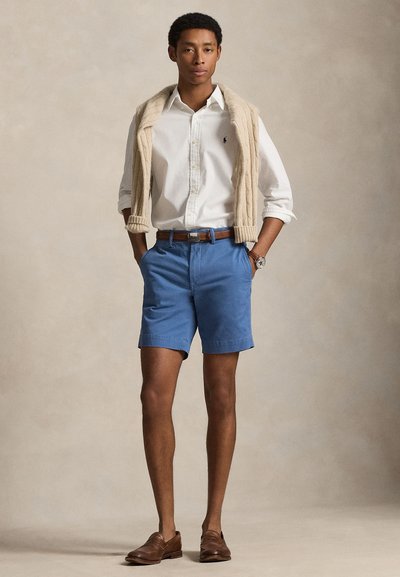 Man draagt een wit overhemd met knopen en een beige trui over zijn schouders, blauwe korte broek, bruine riem, horloge en bruine loafers, staat met handen in de zakken.