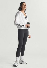 White zip-up jacket with black stripes, black form-fitting leggings, white socks, and gray sneakers. Holding a black cup.
