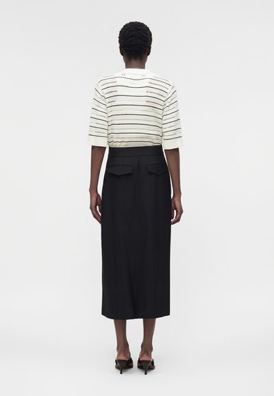 Γυναίκα που φοράει λευκό κοντομάνικο μπλουζάκι με σχέδιο και μαύρη μίντι φούστα με ψηλή μέση και πίσω τσέπες, στέκεται με την πλάτη προς το μέρος μας.