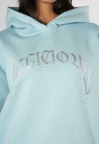 Sudadera azul claro con un estampado gráfico plateado, adornada con joyas de imitación alrededor del texto. Hecha de una tela suave y casual.
