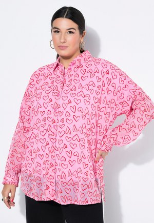 Kvinde med mørkt hår iført en lyserød skjorte med knapper og dækket af røde hjerte-mønstre, står med den ene hånd på hoften.