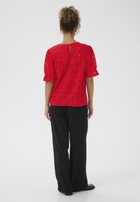 Top en dentelle rouge avec découpes florales, manches bouffantes courtes et dos en trou de serrure ; associé à un pantalon large noir et des chaussures plates.