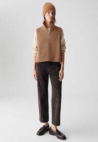 Beige gerippter ärmelloser Pullover mit halbem Reißverschluss, über einem cremefarbenen Langarmshirt getragen, kombiniert mit dunkelbraunen Hosen und braunen Slippern.