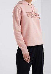 Sudadera rosa claro con un logo brillante en el centro, puños acanalados y capucha con cordón, combinada con pantalones negros. Material de algodón suave.