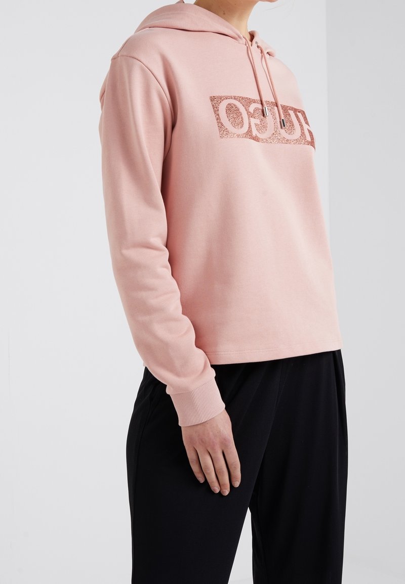 Sudadera rosa claro con un logo brillante en el centro, puños acanalados y capucha con cordón, combinada con pantalones negros. Material de algodón suave.