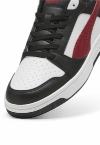 Černobílé tenisky s texturovaným koženým svrškem, červenými akcenty, kulatou špičkou, perforovanou špičkovou částí a bílou gumovou podrážkou.