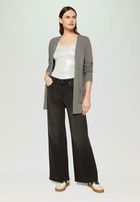 Grijze lange cardigan over een zilveren tanktop, gecombineerd met zwarte wijde broek en beige sneakers. Bevat zakken op de cardigan.