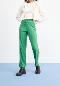 Pantaloni della tuta verdi con strisce bianche laterali, realizzati in un tessuto morbido. Abbinati a una felpa cropped color crema e stivali military neri.