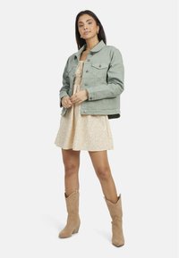 Veste en denim vert clair avec deux poches poitrine, portée sur une robe à motifs floraux. Le mannequin porte également des bottes de cowboy beiges.