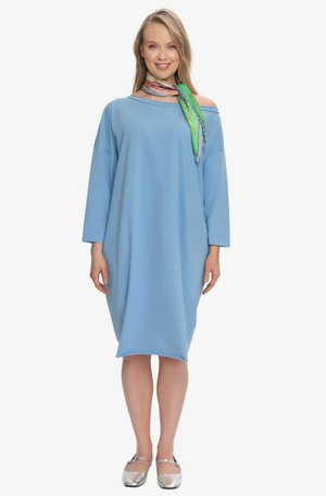 Kobieta ubrana w luźną jasnoniebieską sukienkę odsłaniającą ramiona, wielobarwny szalik i srebrne baleriny, stoi i uśmiecha się na białym tle.