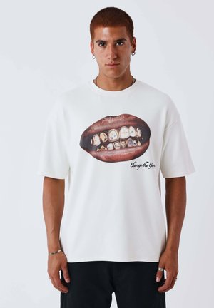 Jonge man met kort haar, gekleed in een wit T-shirt met een grote print van lippen met gouden grills en de tekst "Change the Game".