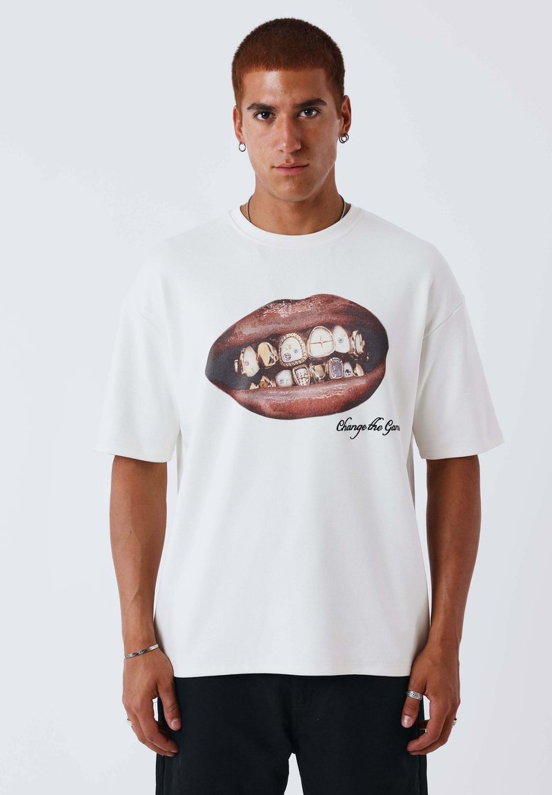 Jonge man met kort haar, gekleed in een wit T-shirt met een grote print van lippen met gouden grills en de tekst "Change the Game".