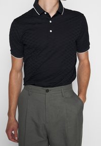 Man som bär en svart strukturerad polotröja med vit kantning och grå högmidjade byxor, med ena handen i fickan, mot en vit bakgrund.