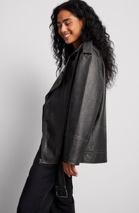 Schwarze Kunstlederjacke mit einem leicht oversized Schnitt, großen Revers und Nähdetails. Verfügt über einen seitlichen Reißverschluss und einen breiten Gürtel.