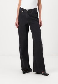 Jeans de mezclilla negros de pierna ancha con cintura alta, costuras en contraste y dobladillo deshilachado. Combinados con zapatos negros de punta afilada.