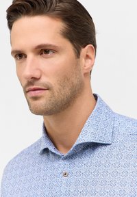 Lichtblauwe shirt met een patroontextuur met geometrische ontwerpen in donkerblauw en oranje. Voorzien van een standaard kraag en knoopsluiting.
