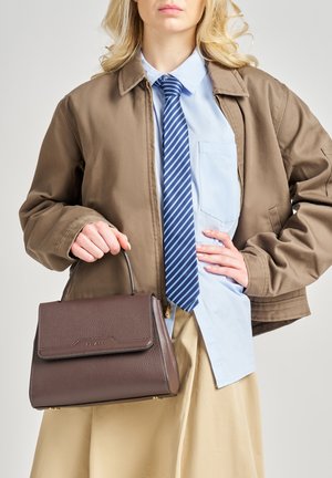 Borsa a mano in pelle marrone con design strutturato, manico superiore e pattina frontale. Il modello indossa una gonna beige, giacca marrone e cravatta a righe blu.