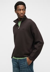 Νεαρός άνδρας με κοντά, σγουρά μαλλιά, φορώντας μαύρο πουλόβερ με φερμουάρ πάνω από πράσινο πουκάμισο και μπλε τζιν, με τα χέρια στις τσέπες, κοιτάζει προς τα αριστερά.