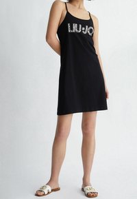 Czarna sukienka bez rękawów z logo "LIU-JO" w wielokolorowych zdobieniach. Modelka ma na sobie białe sandały z złotymi akcentami.