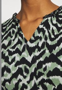 Grün und schwarz gemusterte Bluse mit V-Ausschnitt und Rüschensaum, hergestellt aus strukturiertem Stoff mit wellenförmigem Design.