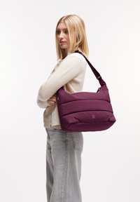 Bolso de hombro morado con un diseño acolchado, textura suave y correa ajustable. Presenta paneles horizontales cosidos y un logo en el lateral.